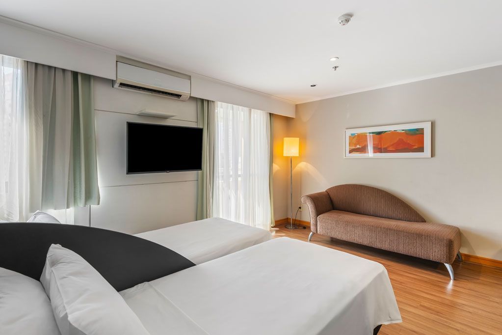 Quarto de hotel moderno com duas camas, TV de parede, sofá e luminária de chão.