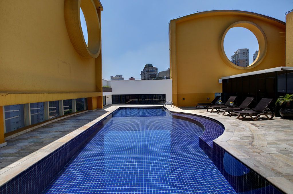 Pátio moderno com piscina, azulejos azuis na água, paredes amarelas e espreguiçadeiras sob um céu claro.