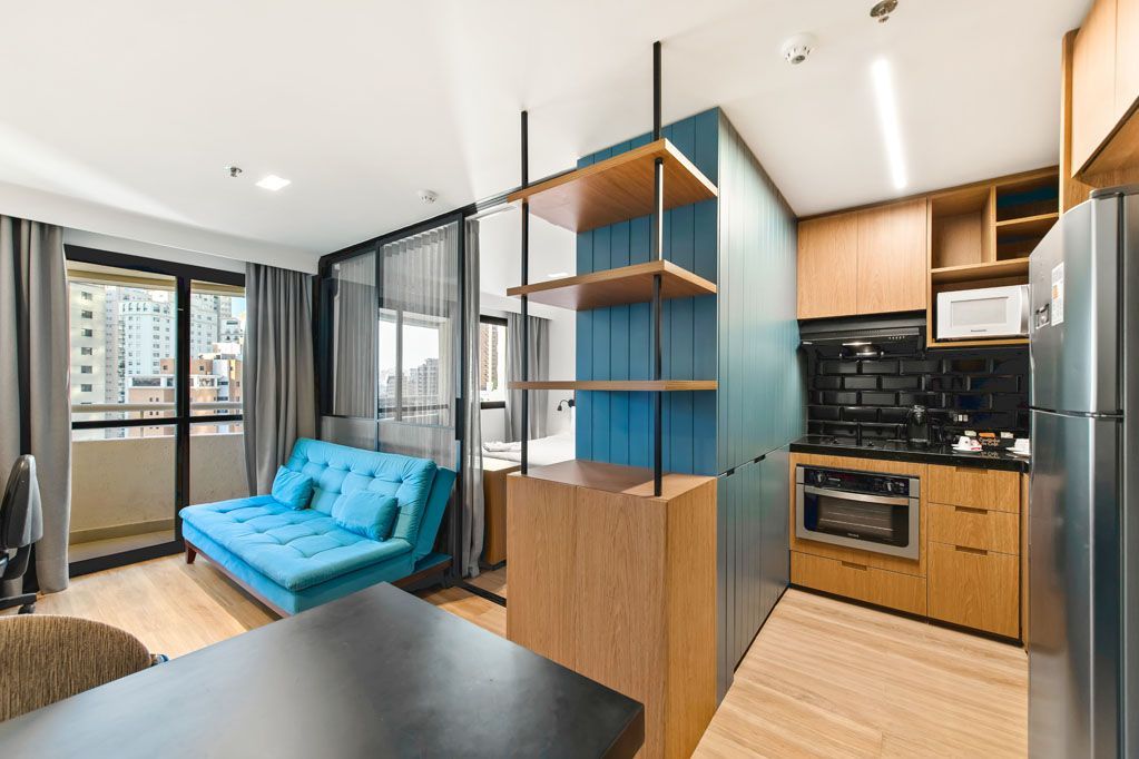 Apartamento estúdio moderno com sofá azul, estantes de madeira e cozinha compacta.