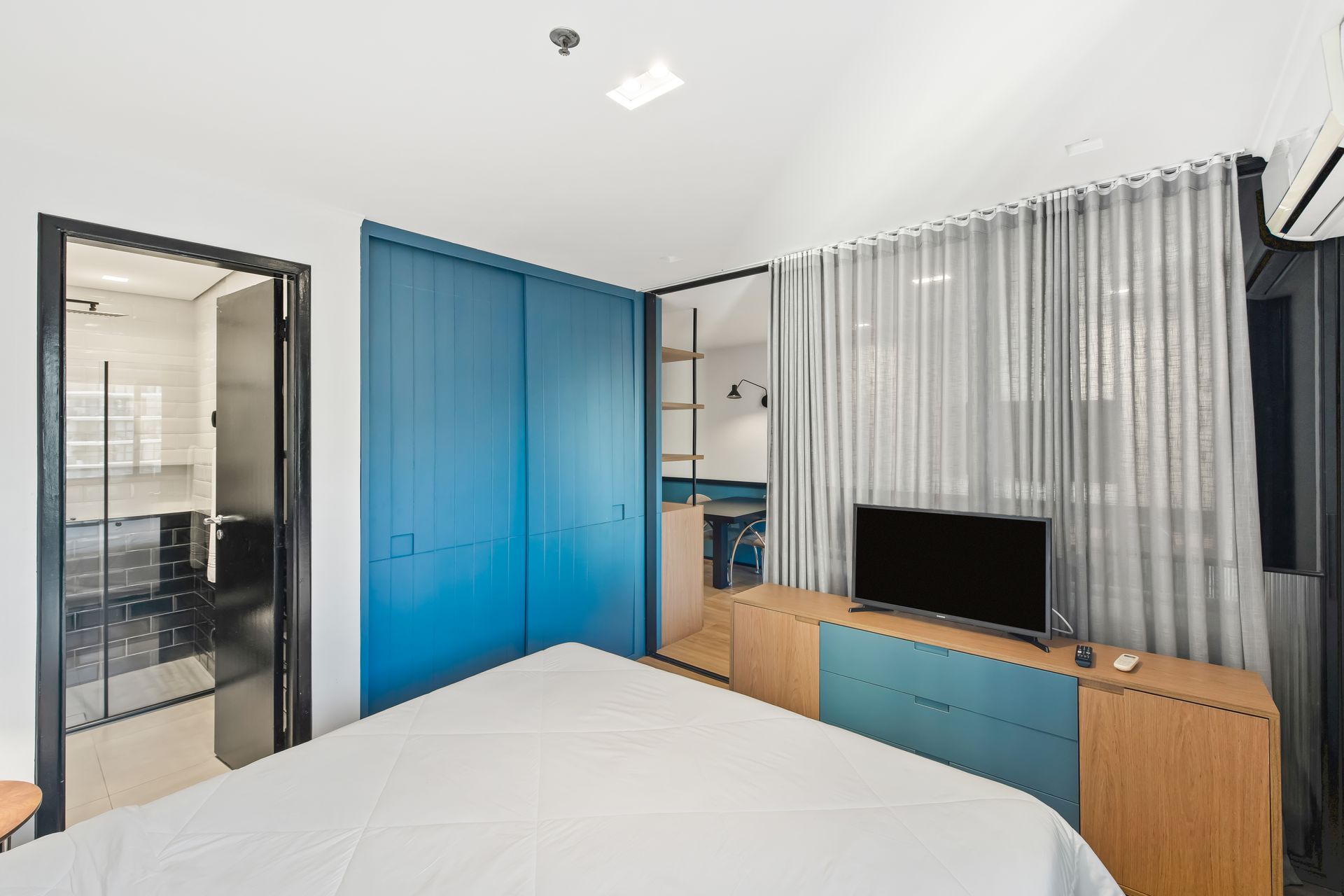 Quarto moderno com guarda-roupa azul, porta de vidro com moldura preta, TV e cortinas translúcidas.