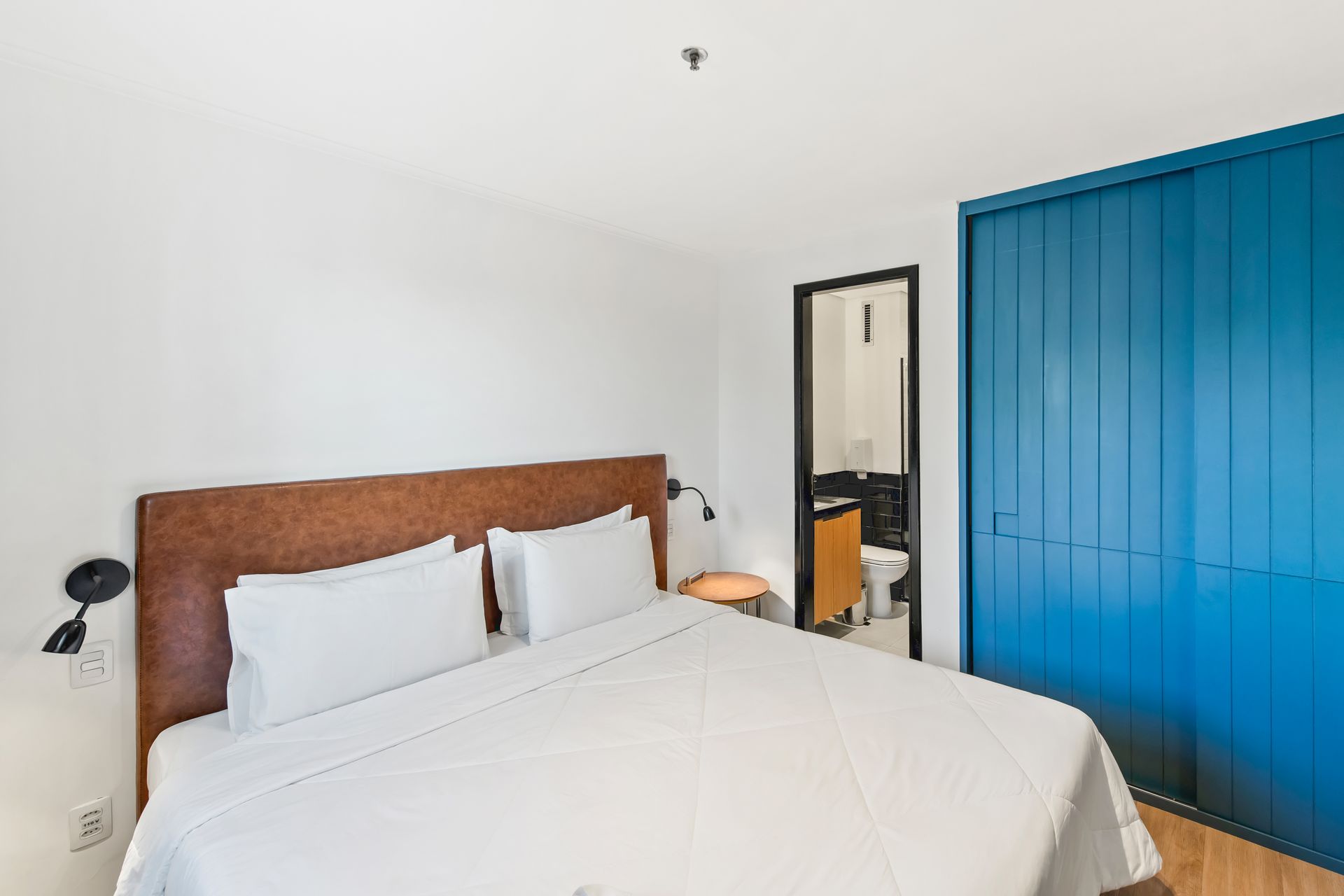 Quarto luminoso com cama, guarda-roupa azul e espelho em parede branca.