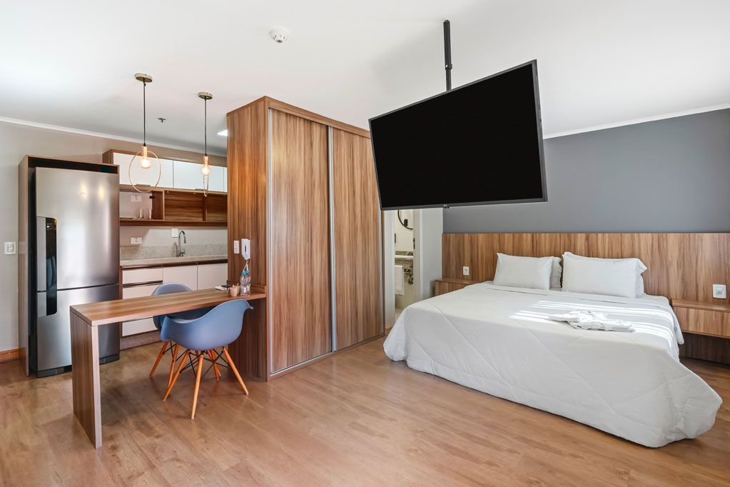Apartamento estúdio moderno com cama, escrivaninha, divisória de madeira e área de cozinha.