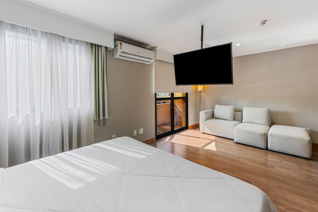Quarto iluminado com cama, sofá branco, janela grande e TV de parede com luz quente.
