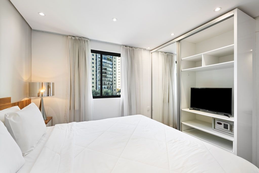 Quarto moderno e luminoso, todo branco, com uma cama grande, janela e prateleiras embutidas para a TV.