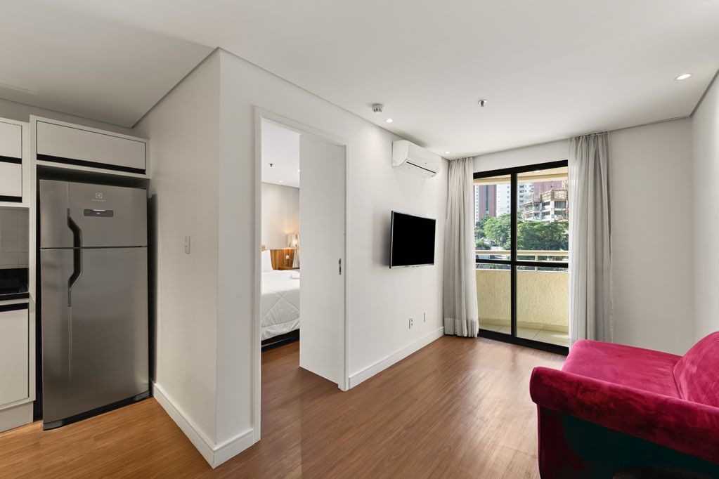 Apartamento moderno e luminoso com cozinha, TV de parede, portas de varanda e sofá vermelho sobre piso de madeira.