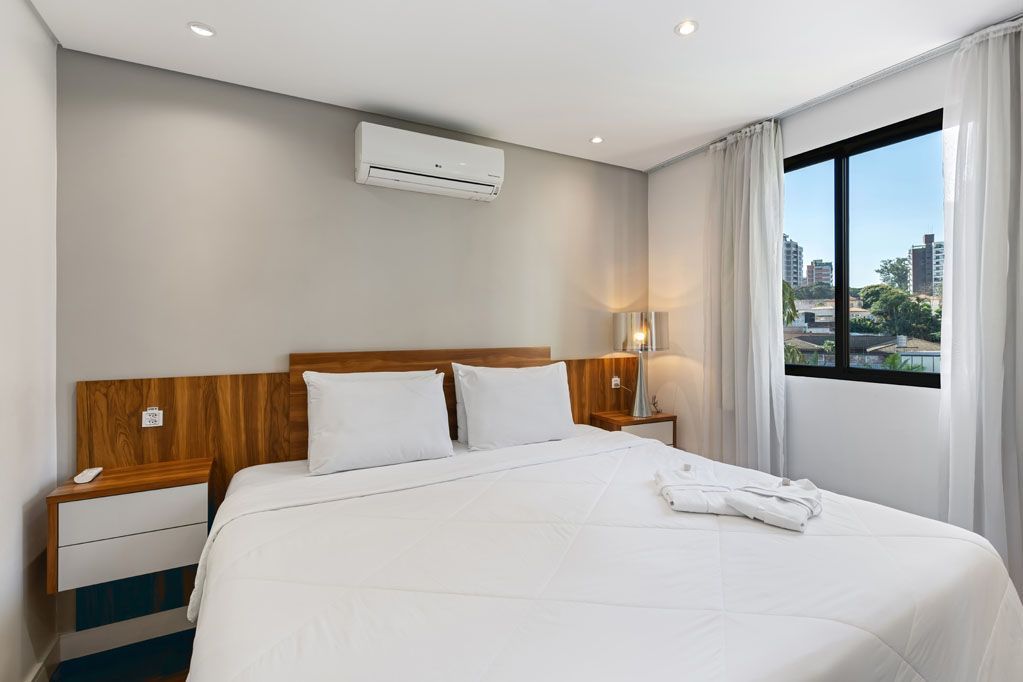 Quarto moderno e luminoso com cama branca, cabeceira de madeira, candeeiros de cabeceira e janela com vista para a cidade.
