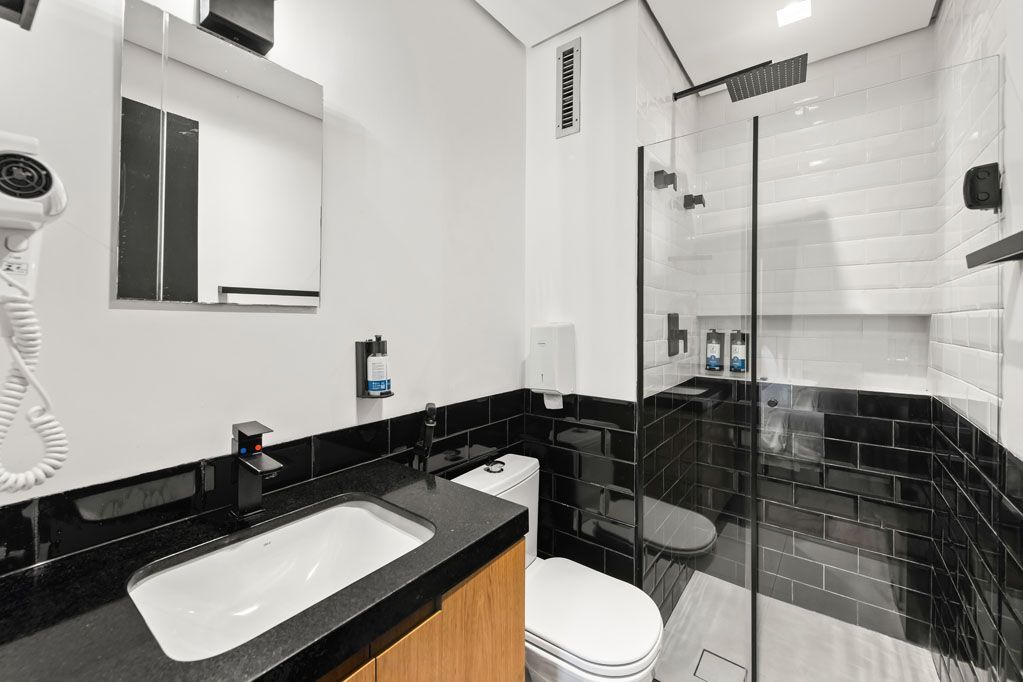 Banheiro moderno com bancadas pretas, azulejos brancos, pia, vaso sanitário, espelho e box de vidro.