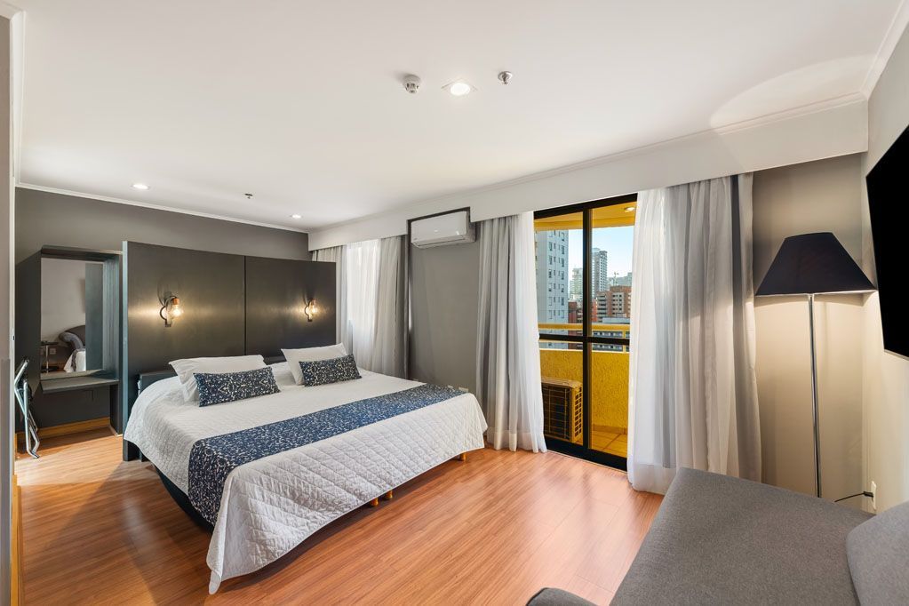 Quarto de hotel moderno com cama, luminária de chão, guarda-roupa amplo e portas de varanda com vista para a cidade.