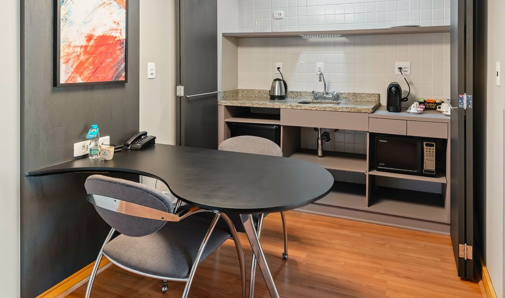 Cantinho de cozinha moderno com mesa redonda preta, cadeiras e uma bancada compacta com pia e chaleira.