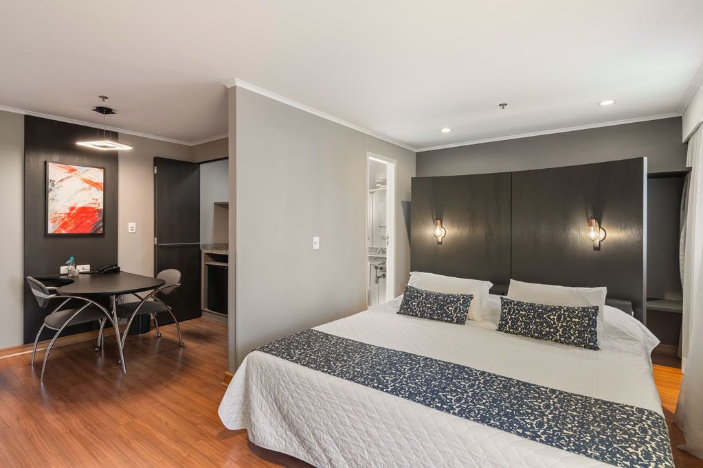Quarto moderno com paredes cinza, cabeceira escura, roupa de cama branca e uma área de jantar com mesa de vidro além da porta aberta.