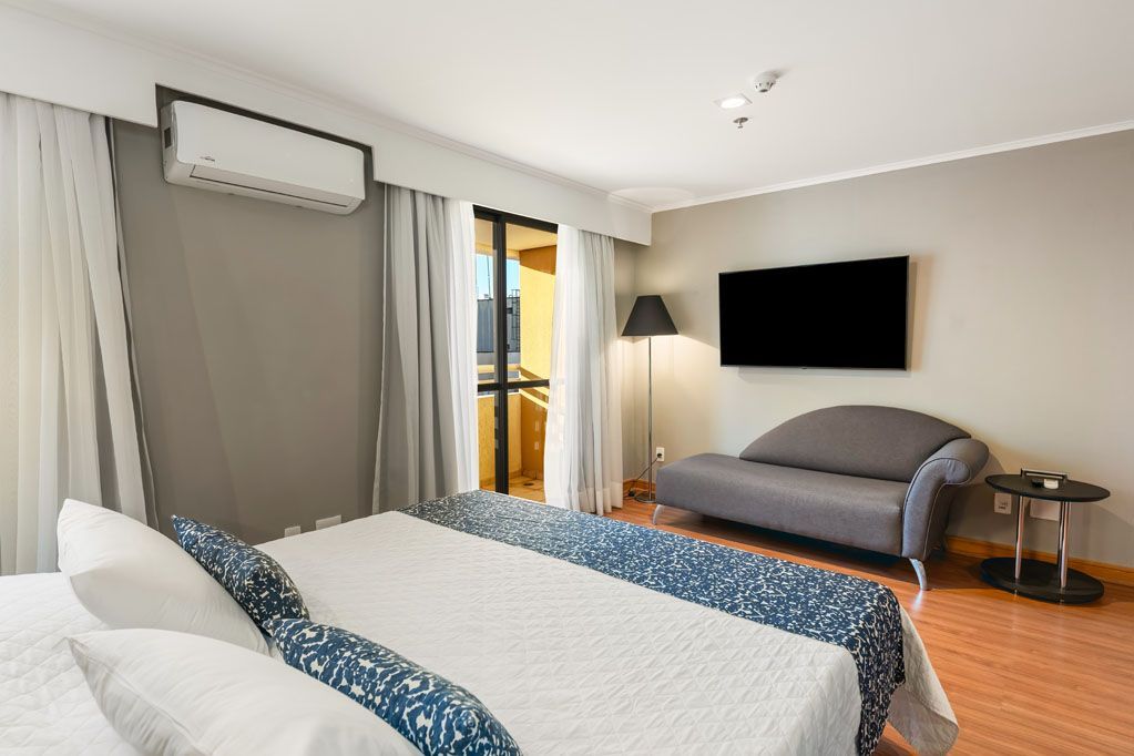 Quarto de hotel com duas camas de solteiro, TV de parede, sofá, porta para varanda e cortinas grandes na janela.
