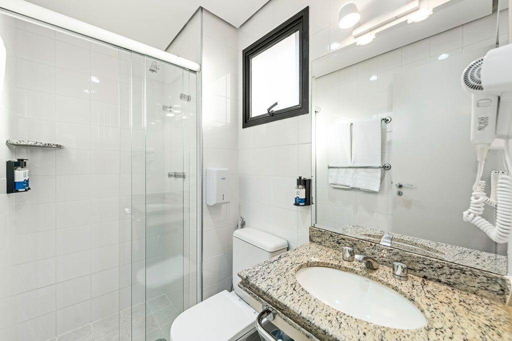 Banheiro branco e luminoso com box de vidro, bancada de granito com pia, espelho e secador de cabelo fixado na parede.