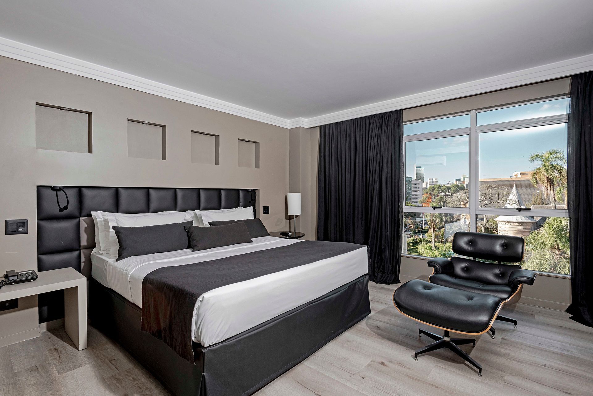 Quarto de hotel moderno com cama grande, poltrona preta e vista da cidade através de uma janela.