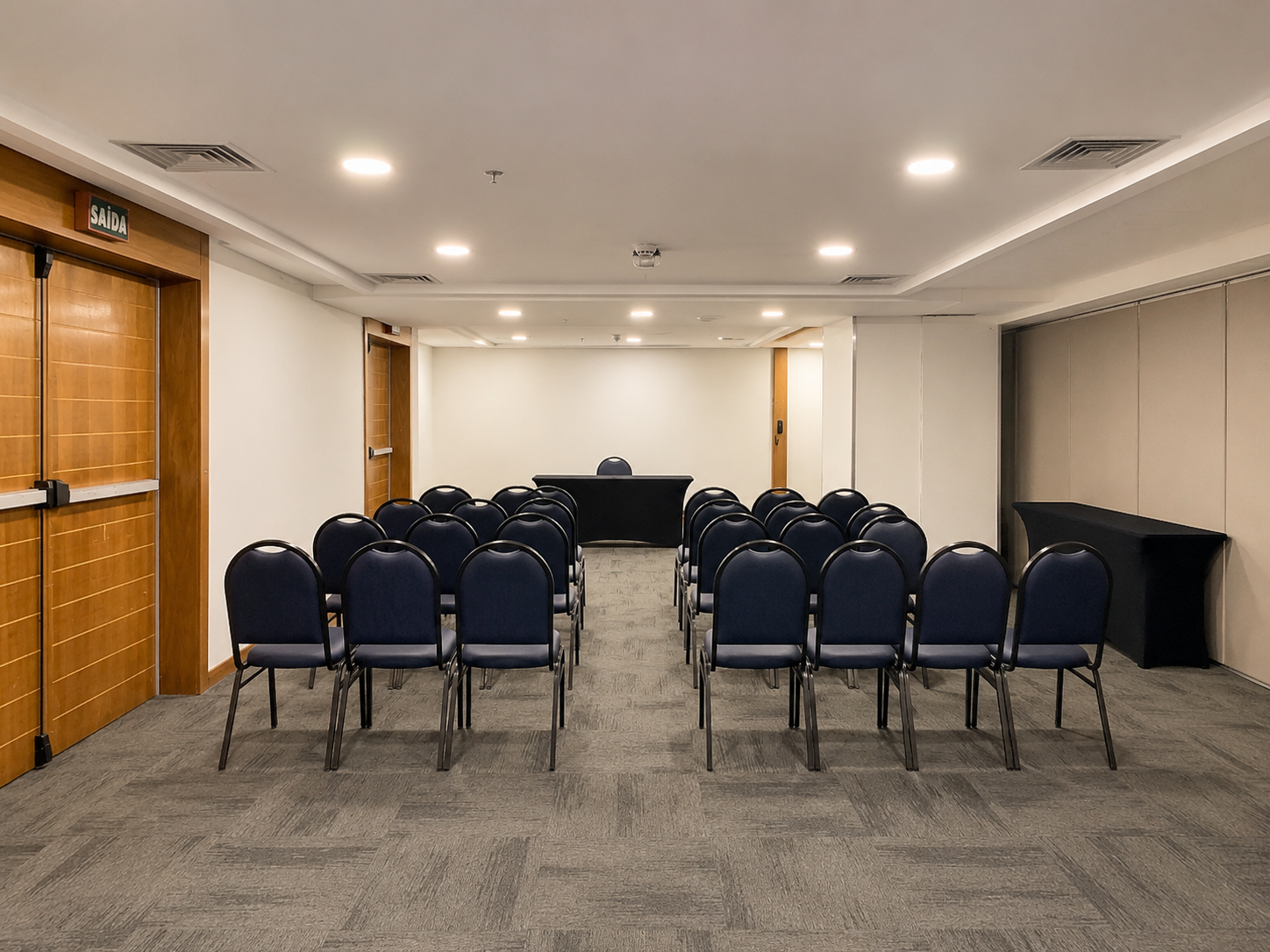 Sala de conferências vazia com fileiras de cadeiras azuis voltadas para uma mesa e uma tela de projeção.