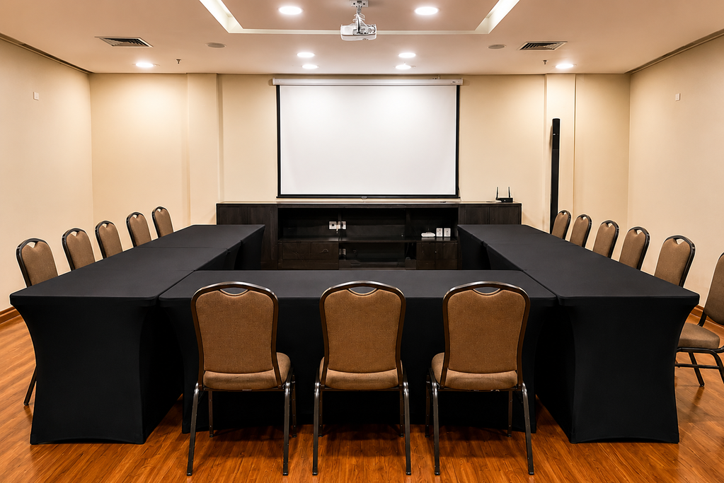 Sala de conferências vazia com mesas pretas em formato de U, cadeiras bege e uma grande tela de projeção na parede frontal.
