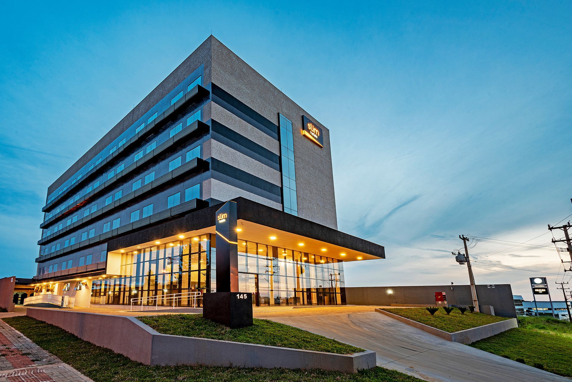 Edifício de escritórios moderno com fachada de vidro ao entardecer, com iluminação aconchegante no hall de entrada e uma rampa de acesso para veículos.