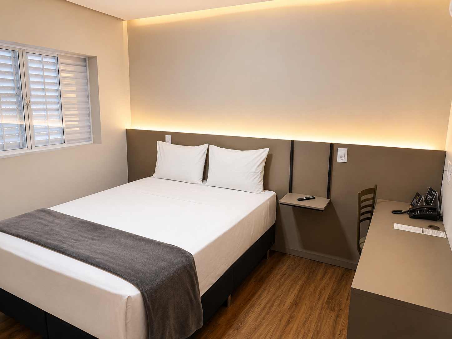 Quarto de hotel minimalista com cama, escrivaninha, cadeira e iluminação ambiente aconchegante.