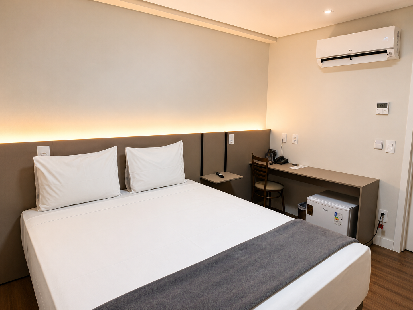Quarto de hotel moderno com cama branca, escrivaninha, cadeira e ar-condicionado de parede.