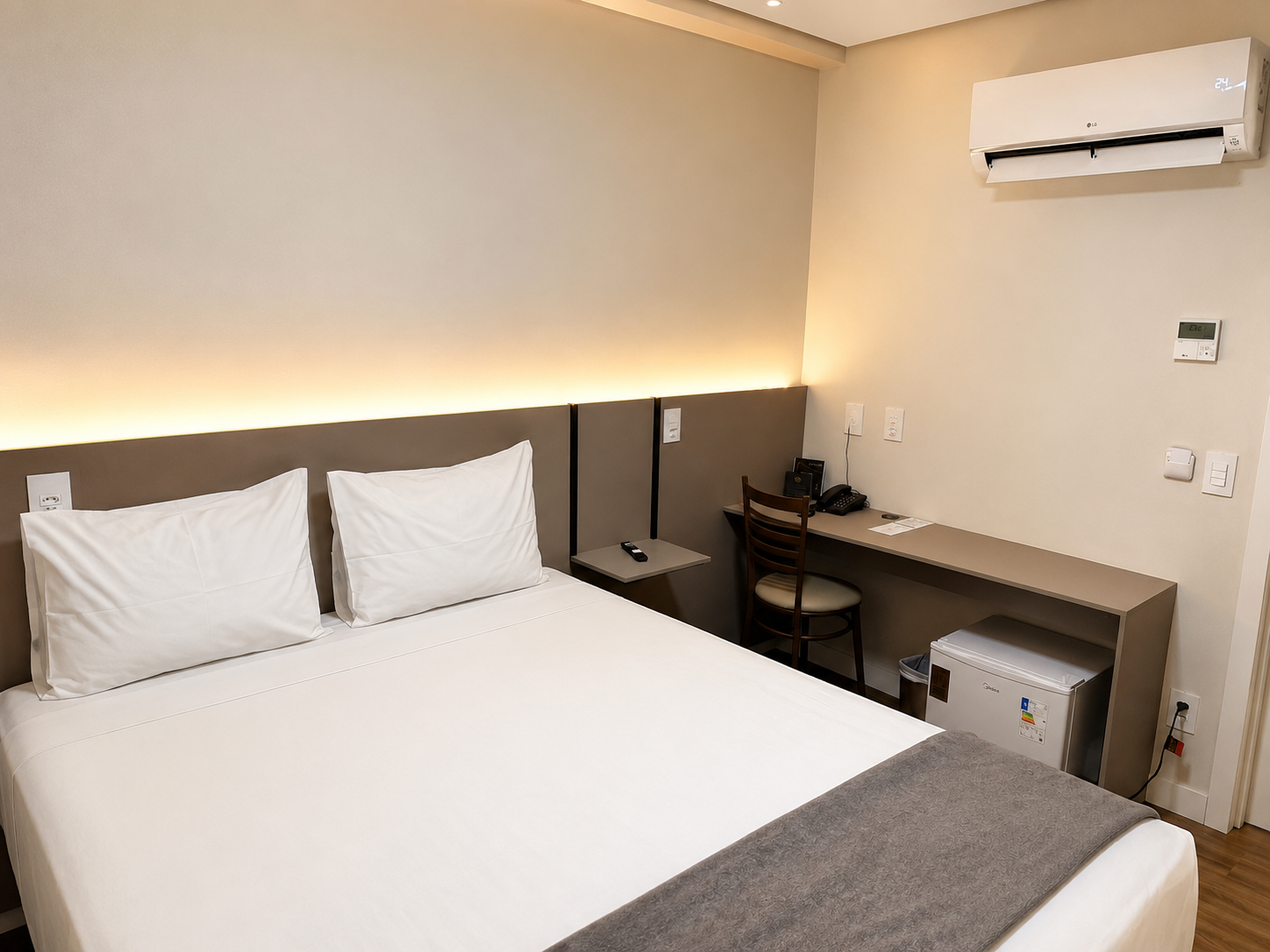 Quarto de hotel com duas camas de solteiro, escrivaninha, cadeira, frigobar e ar-condicionado de parede.
