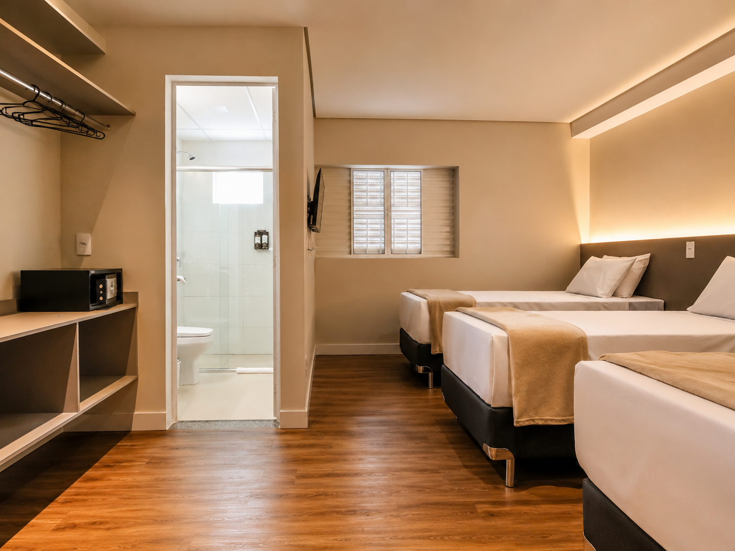 Quarto de hotel moderno com duas camas, piso de madeira em tons quentes, paredes bege e um pequeno banheiro visível através de uma porta.