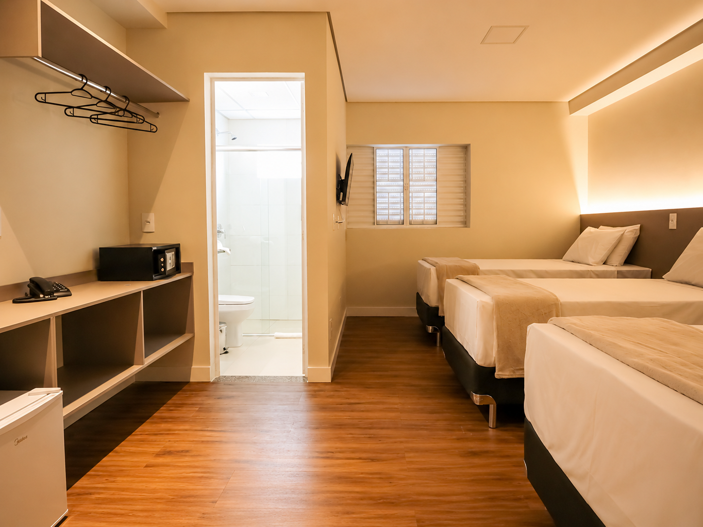 Quarto de hotel simples com duas camas, escrivaninha, armário aberto e porta do banheiro, em tons de bege quente.