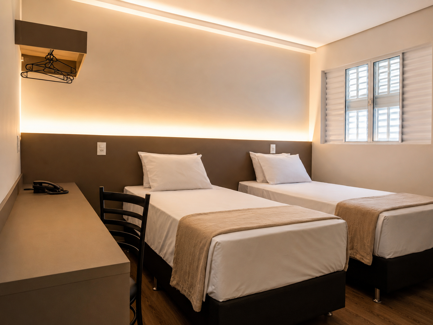 Duas camas de solteiro em um quarto de hotel com iluminação aconchegante, escrivaninha e persianas nas janelas.