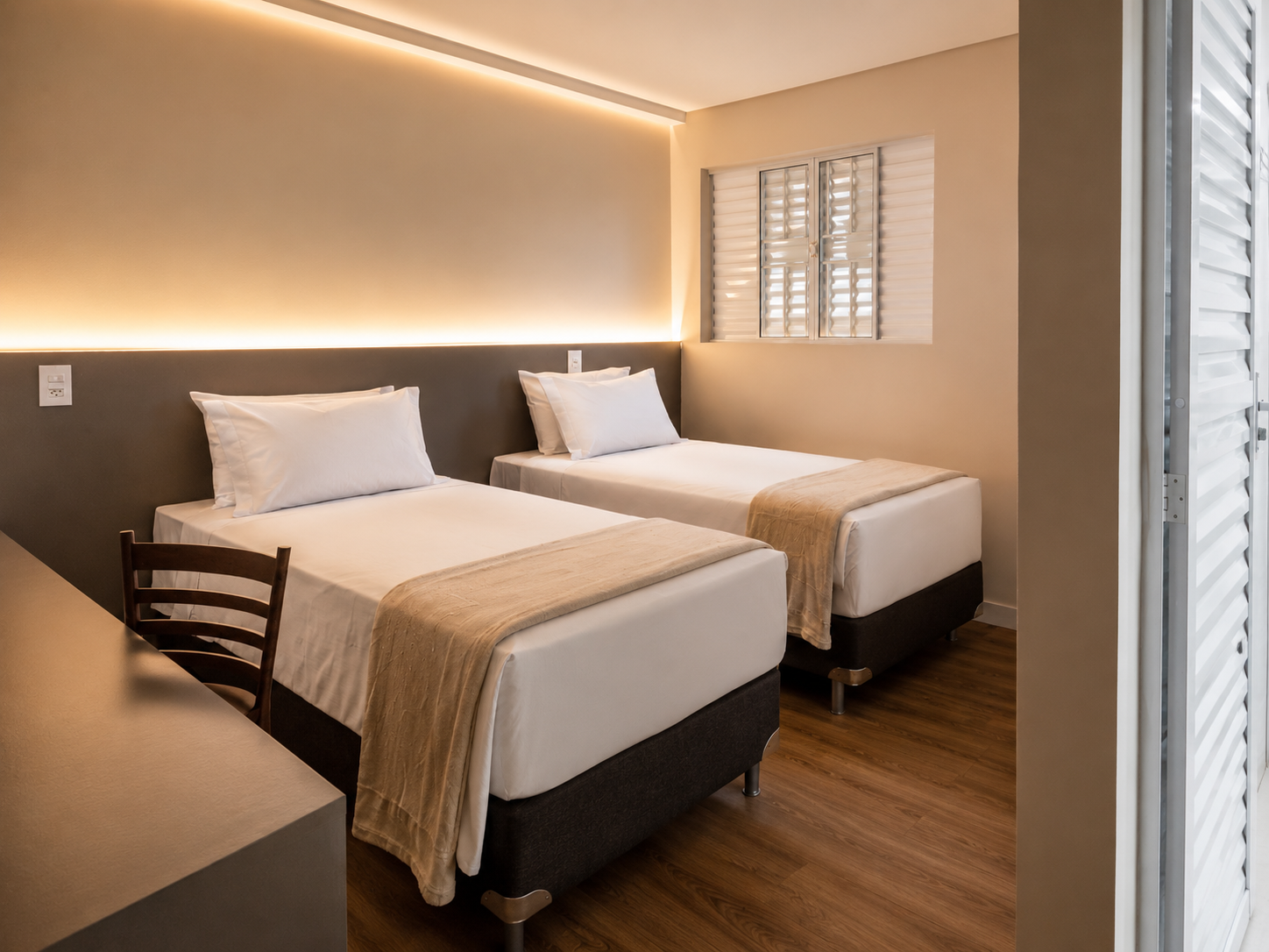 Quarto de hotel pequeno com duas camas de solteiro, paredes bege, piso de madeira e janela com persianas.