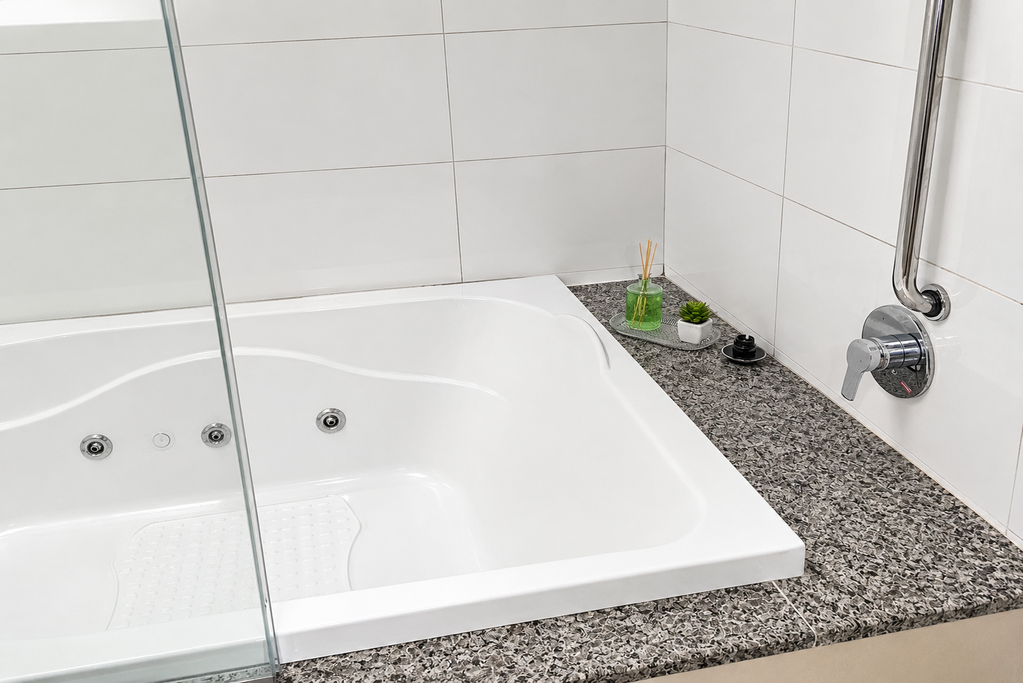 Banheira branca com painel de vidro para o chuveiro ao lado de uma bancada de granito salpicado e parede revestida com azulejos.
