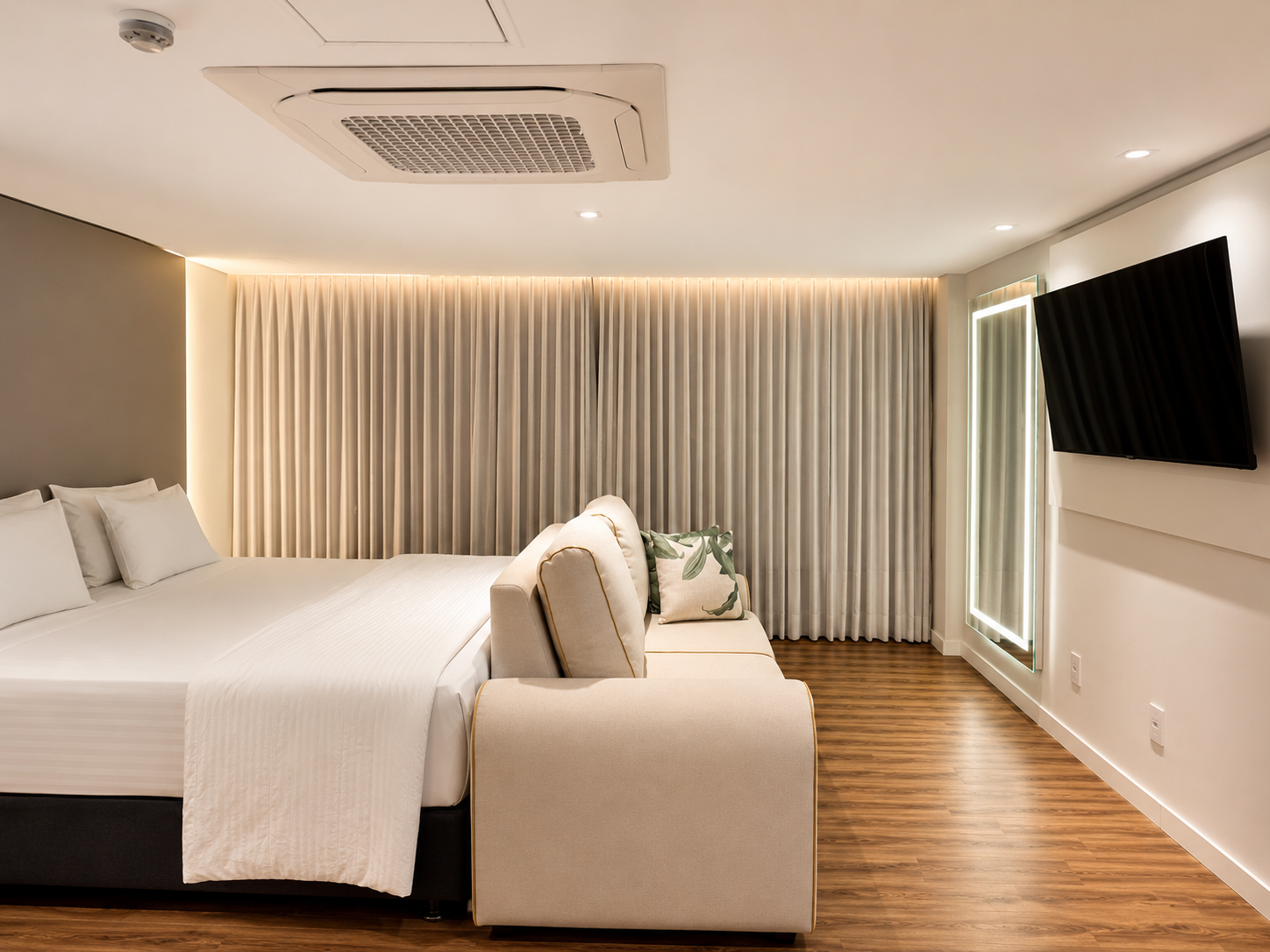 Quarto moderno com cama grande, sofá bege, piso de madeira, ar condicionado de teto e TV de parede.
