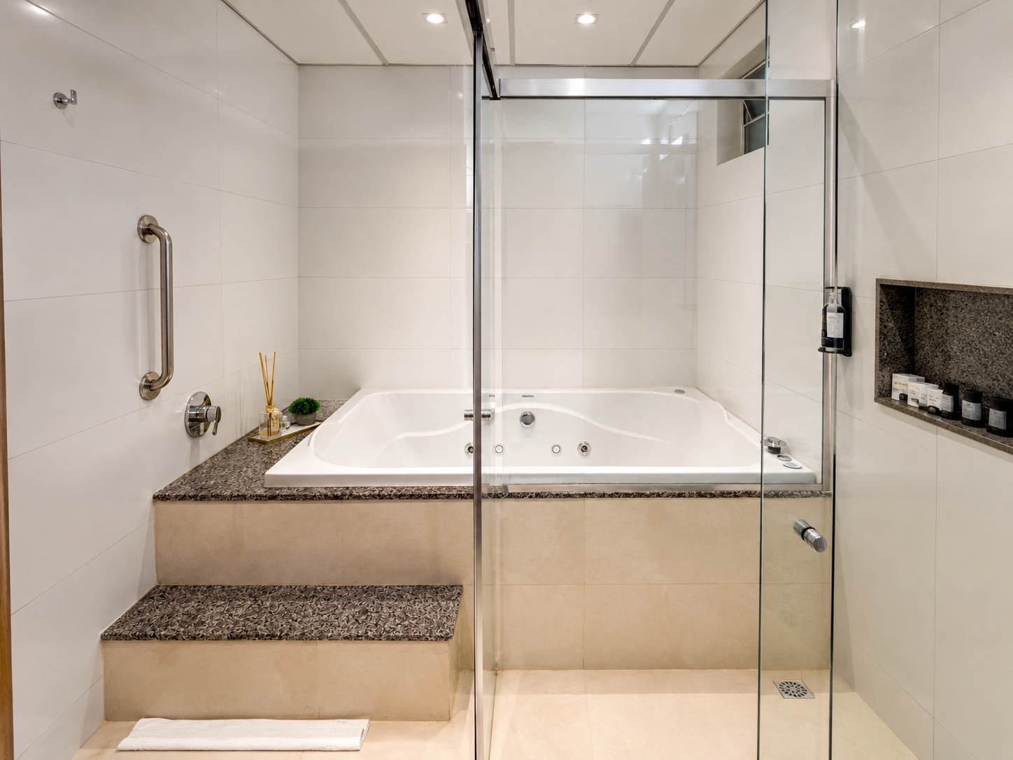 Banheiro moderno com banheira branca elevada atrás de um box de vidro e degraus revestidos com azulejos bege.
