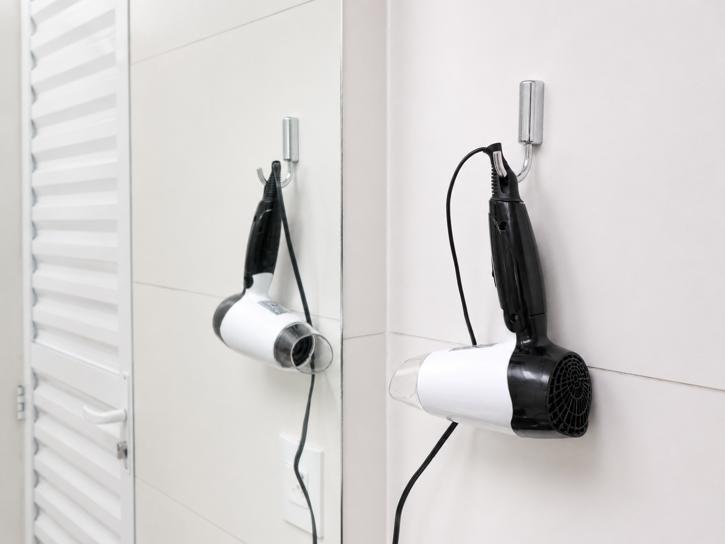 Dois secadores de cabelo fixados em uma parede branca ao lado de uma porta veneziana, com os cabos pendurados.