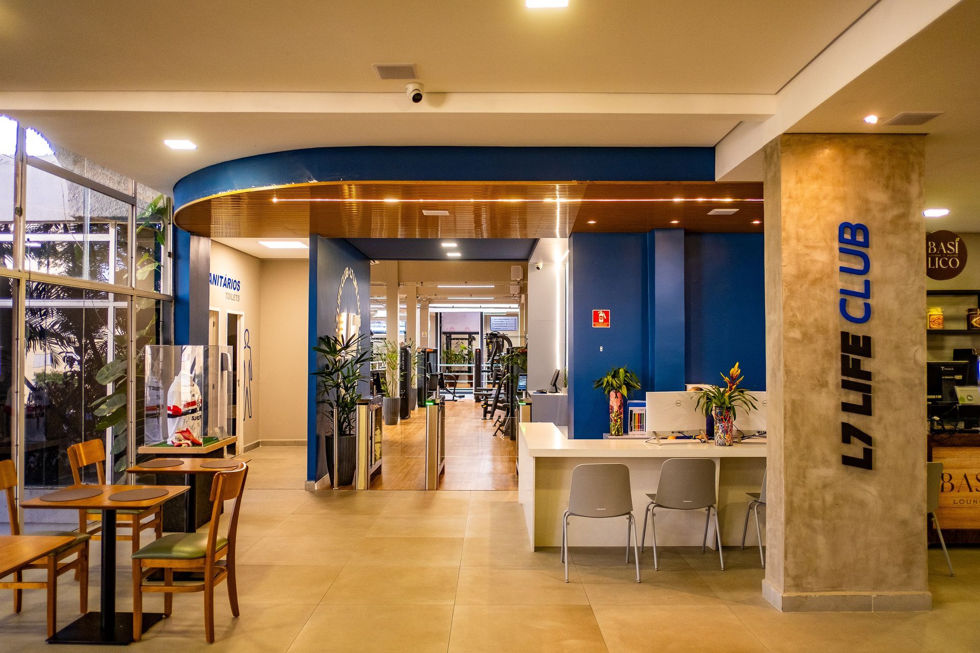 Interior moderno do lobby com paredes azuis, detalhes em madeira, mesas, plantas e um corredor que leva ao interior do edifício.