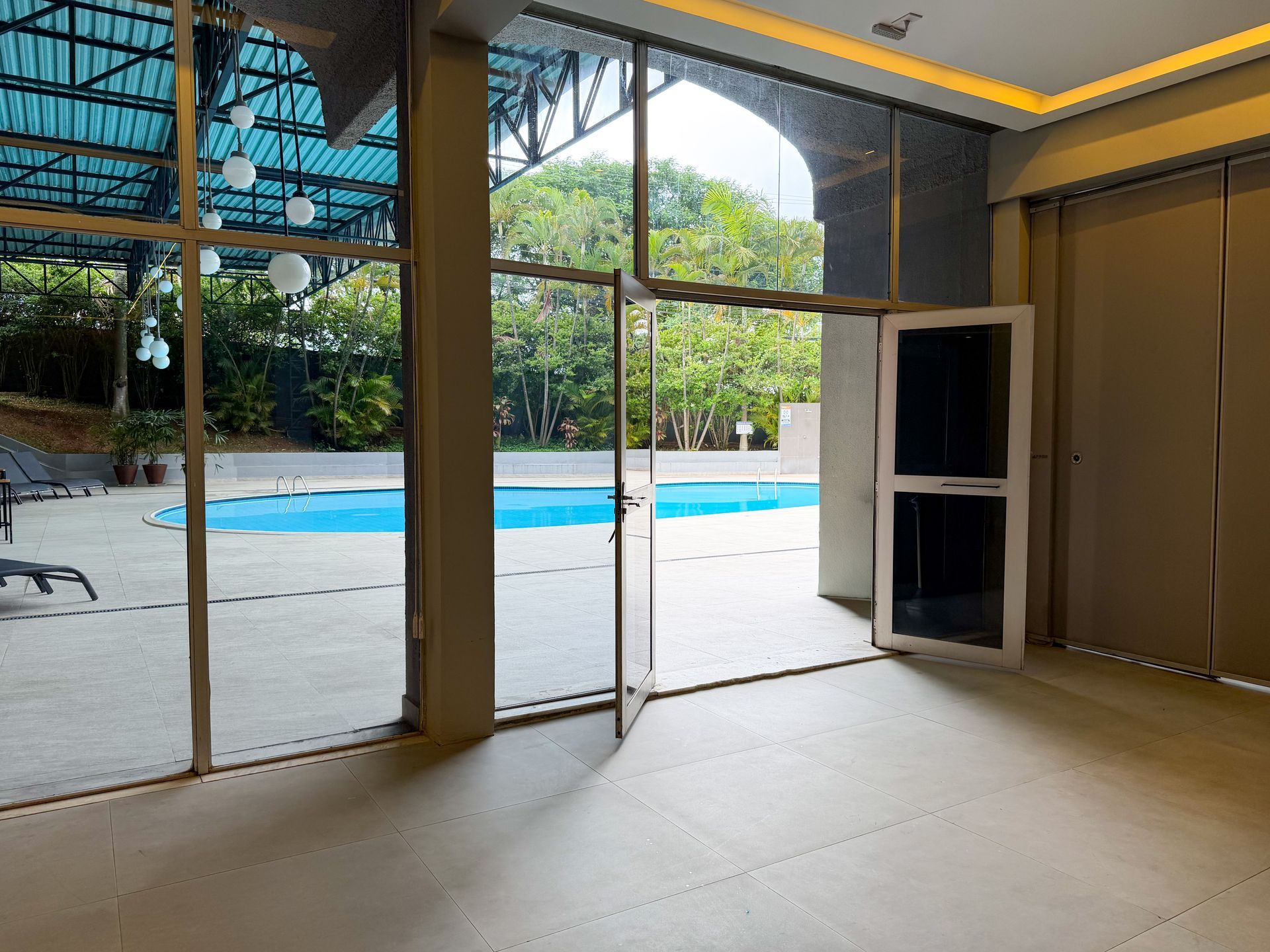 Sala interna vazia com portas de vidro que se abrem para uma piscina e um pátio arborizado.