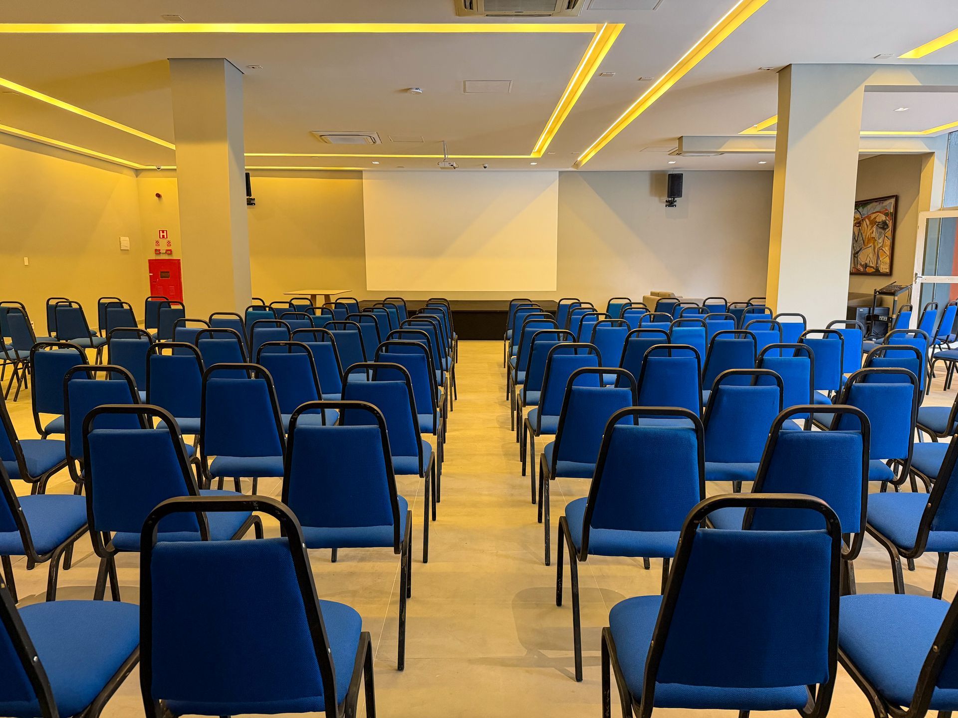 Sala de conferências vazia com fileiras de cadeiras azuis voltadas para um palco e uma tela branca sob luzes quentes no teto.
