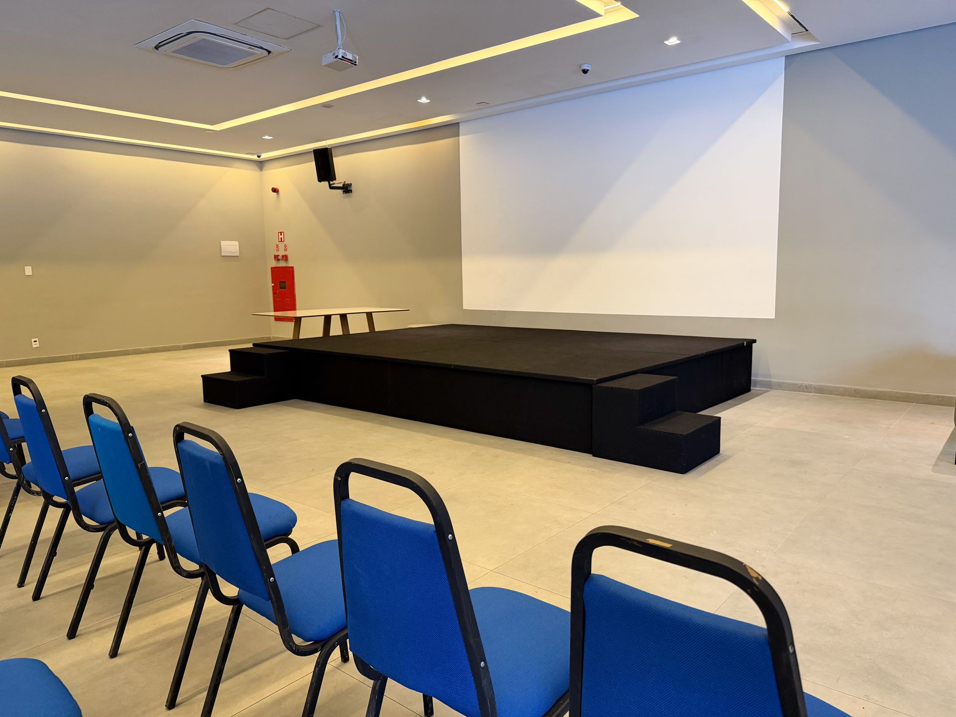 Sala de conferências vazia com cadeiras azuis, palco preto e grande tela de projeção.