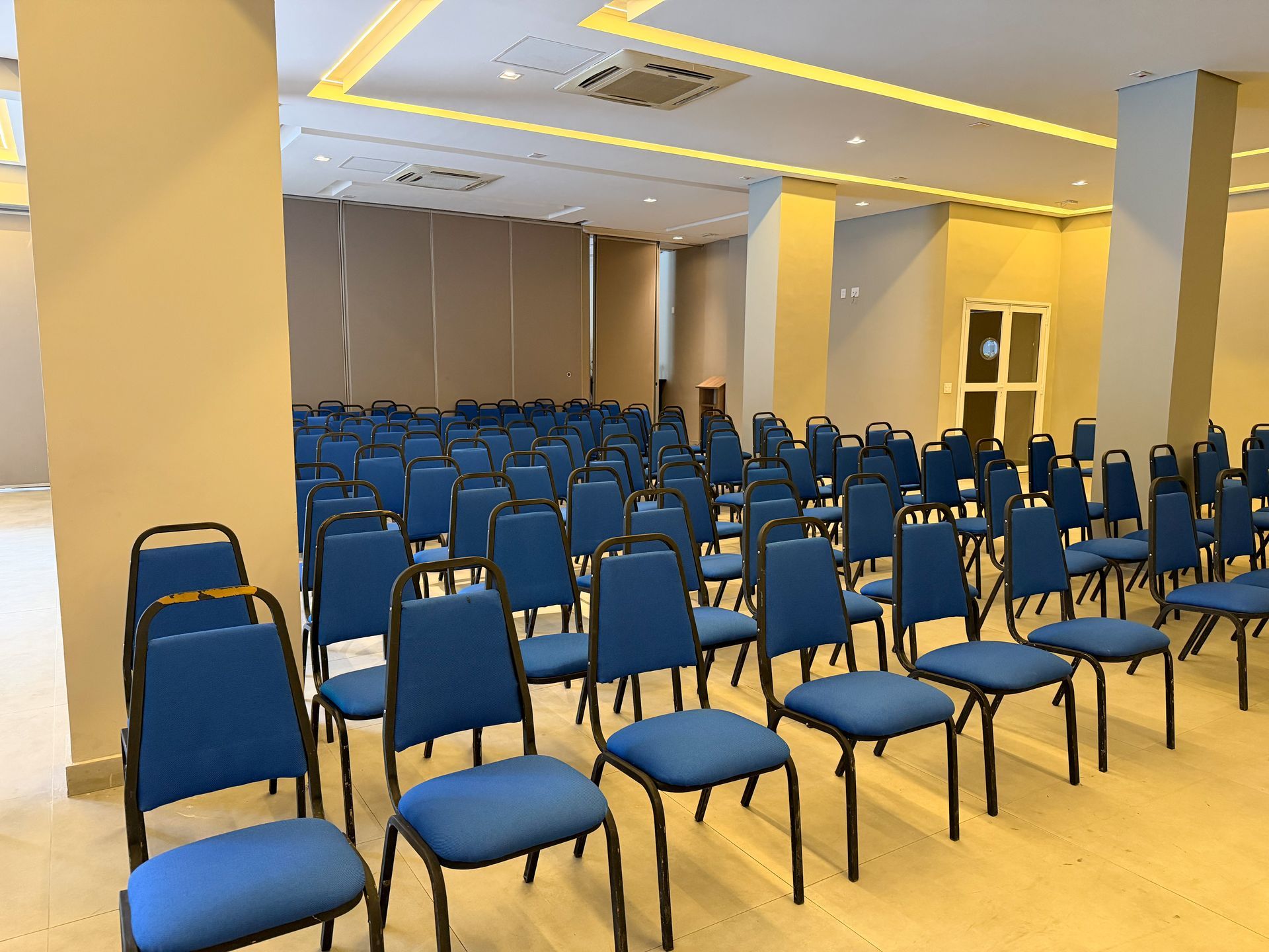 Fileiras de cadeiras azuis vazias em uma sala de conferências com paredes bege e luzes no teto.