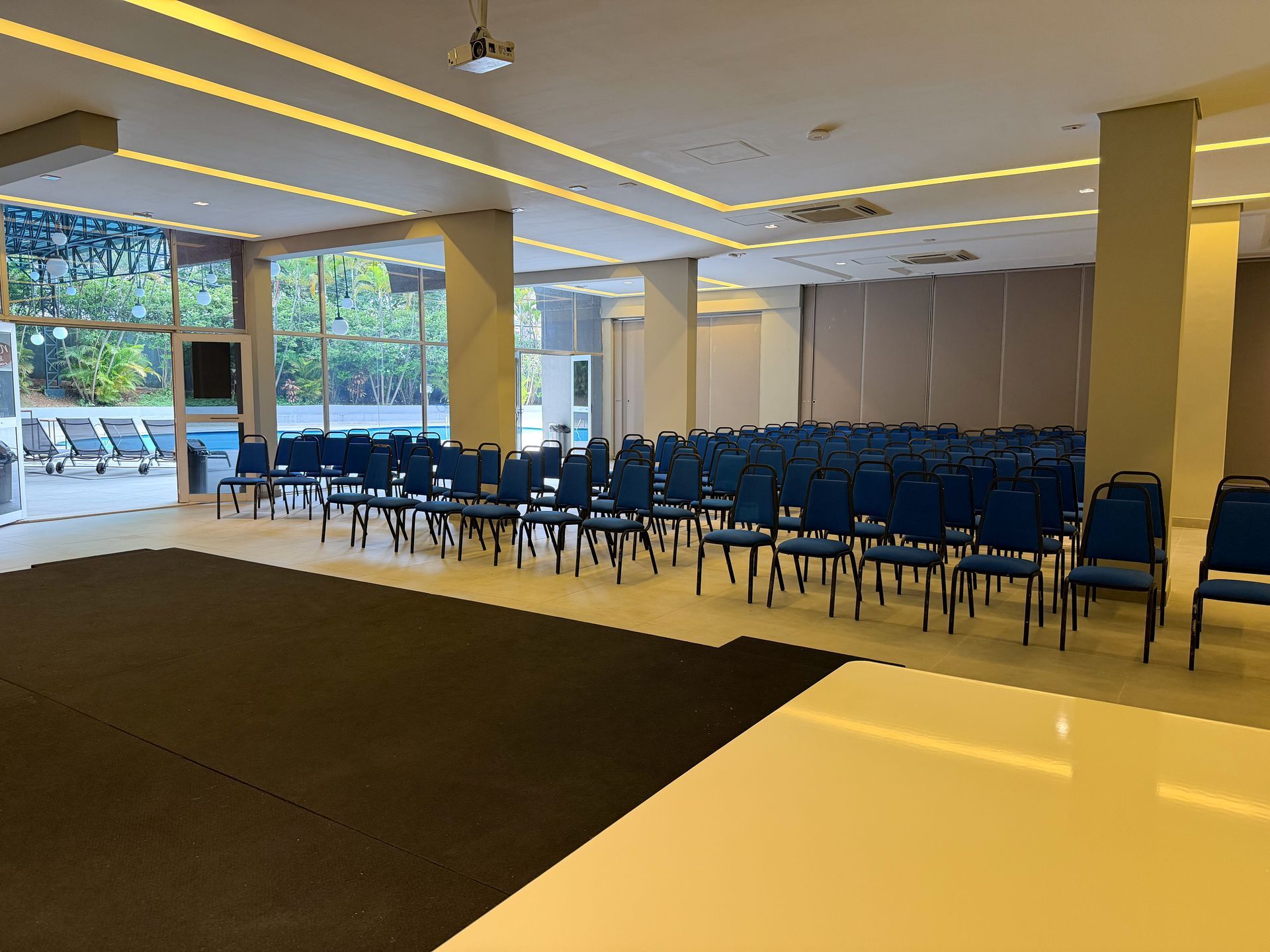 Sala de conferências vazia com fileiras de cadeiras azuis, colunas bege e grandes janelas com vista para as árvores.