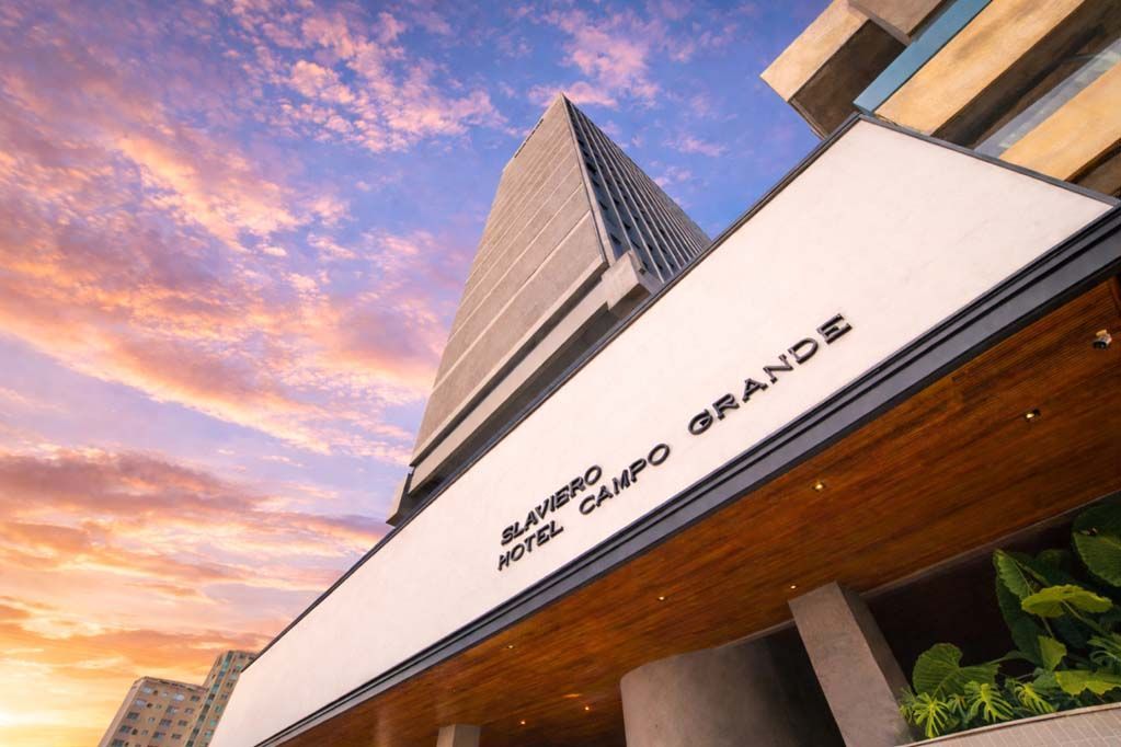 Vista de baixo para cima de um prédio de hotel moderno ao pôr do sol, com céu rosa-azulado e palmeiras.
