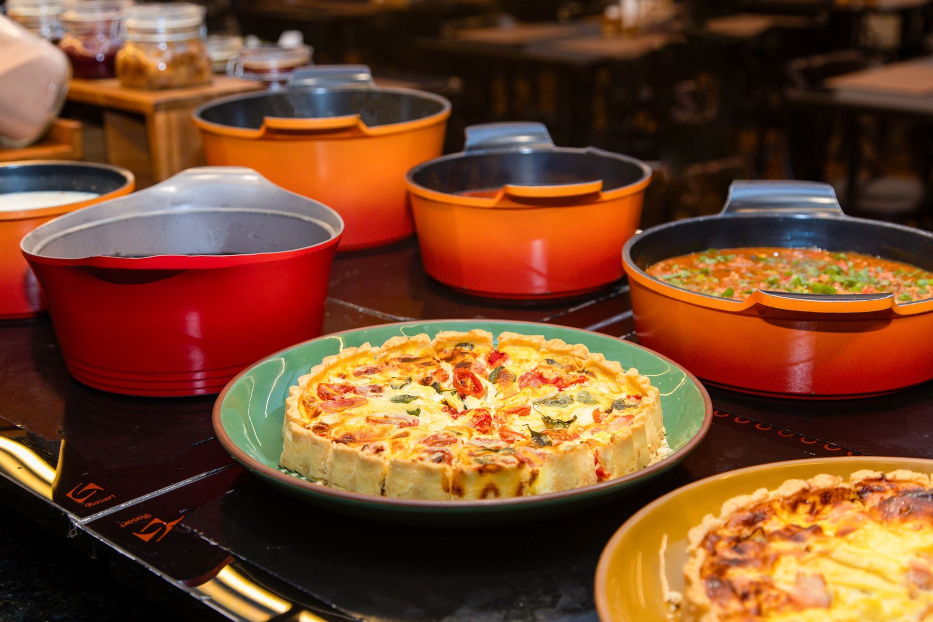 Mesa de bufê com caçarolas e pratos assados variados em assadeiras vermelhas, laranjas e verdes.