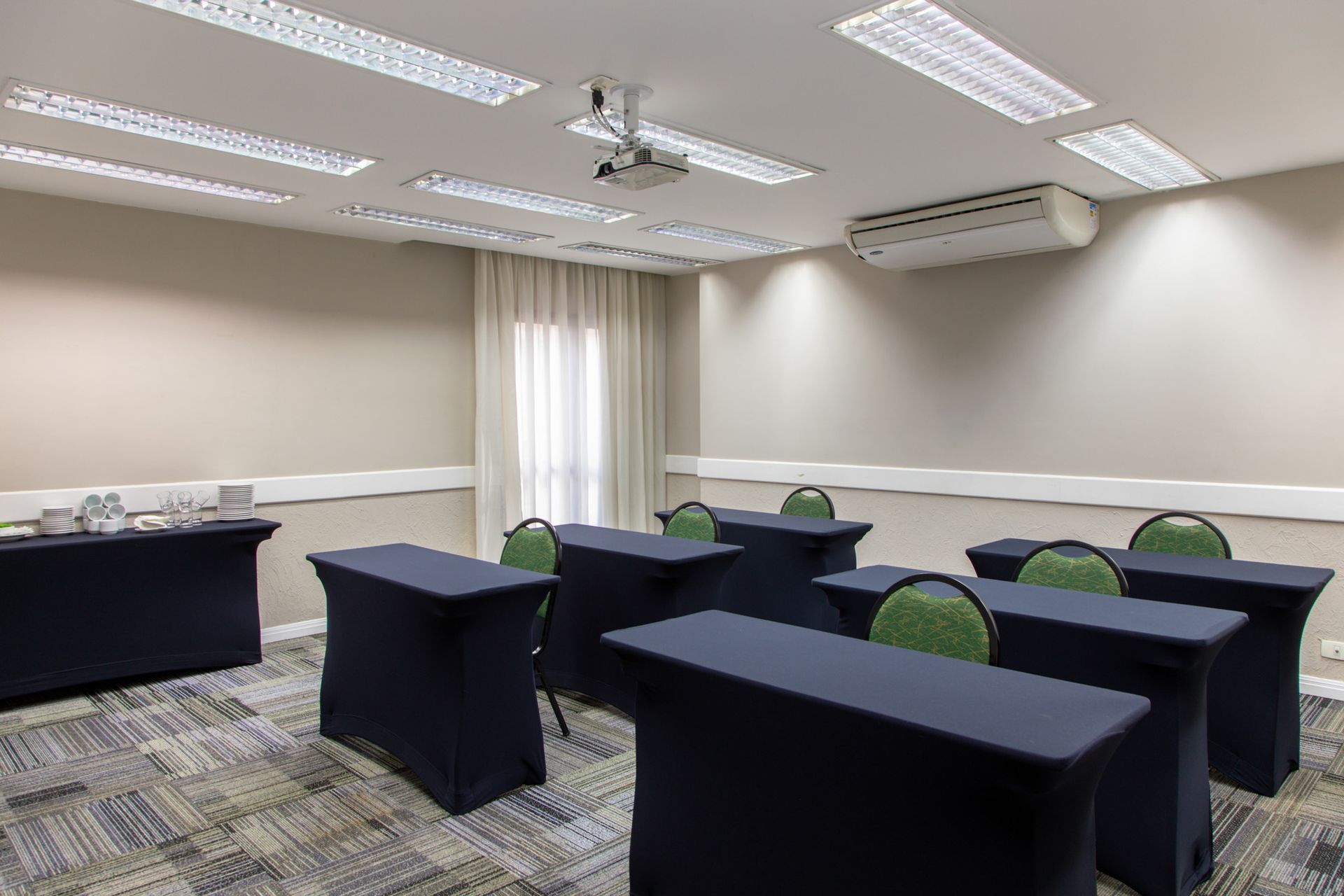 Sala de conferências vazia com mesas escuras, cadeiras verdes e luzes fluorescentes no teto.