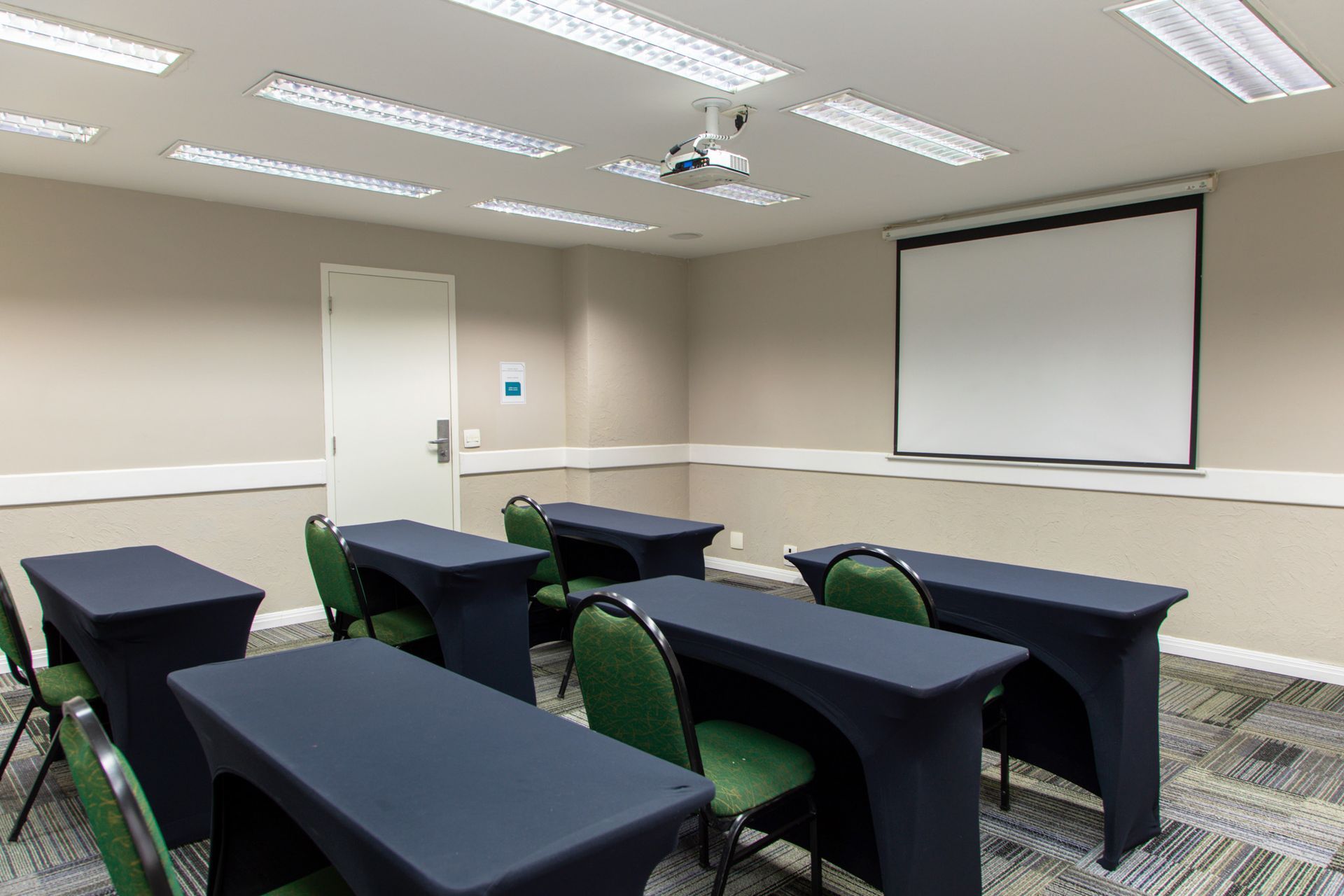Sala de aula vazia com fileiras de mesas pretas e cadeiras verdes, tela de projeção e luzes fluorescentes no teto.