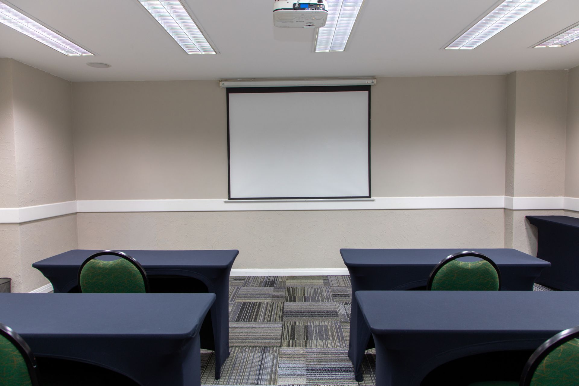 Sala de aula vazia com carteiras voltadas para uma tela de projeção retrátil na parede frontal.