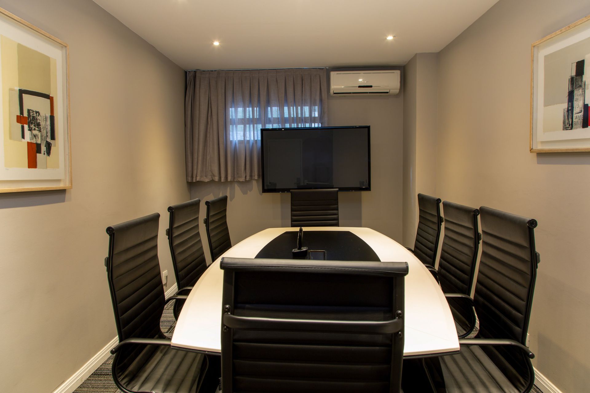 Sala de conferências moderna com cadeiras pretas, mesa branca, obras de arte na parede, TV e ar condicionado.