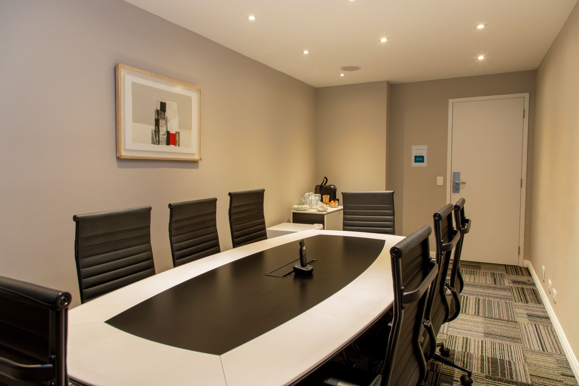Sala de conferências moderna com mesa oval, cadeiras pretas, obras de arte na parede e porta fechada.