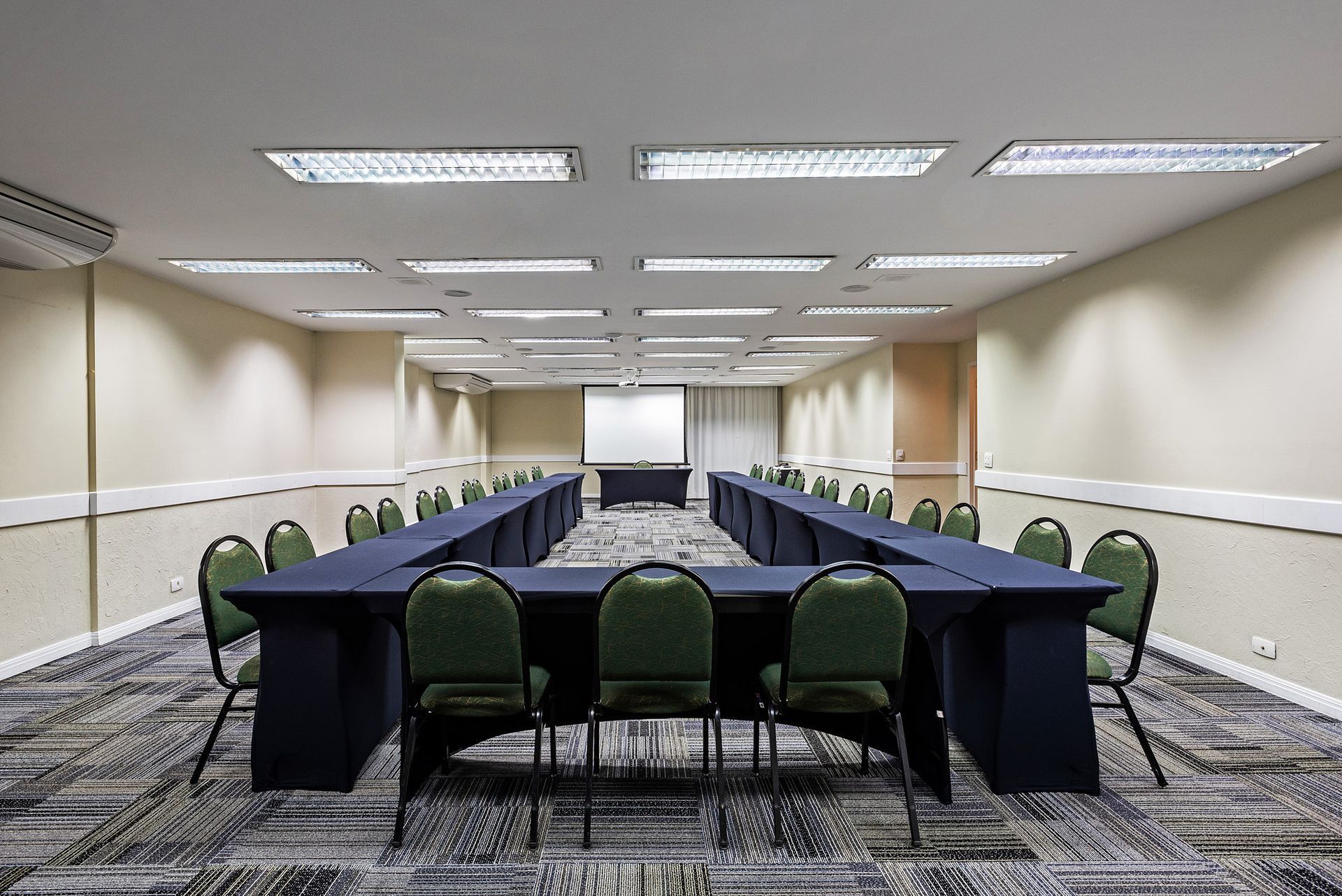 Sala de conferências com mesa comprida em formato de U, cadeiras verdes e luzes fluorescentes no teto.