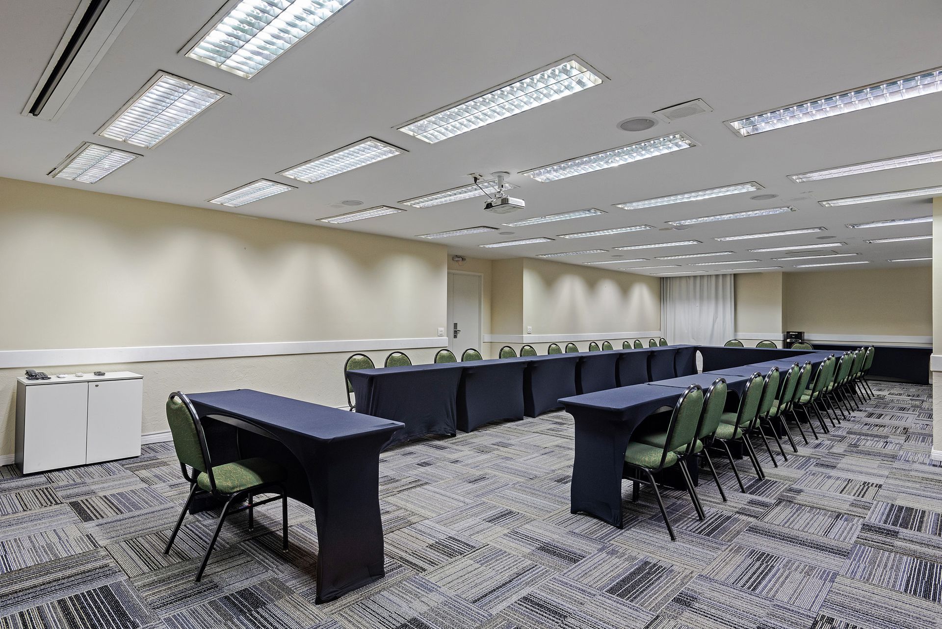 Sala de conferências vazia com mesas compridas, cadeiras verdes e luzes fluorescentes brilhantes no teto.