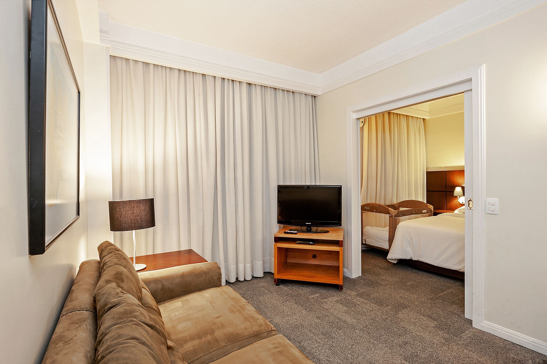 Sala de estar do quarto de hotel com sofá, TV em suporte de madeira e porta para um quarto com cama.