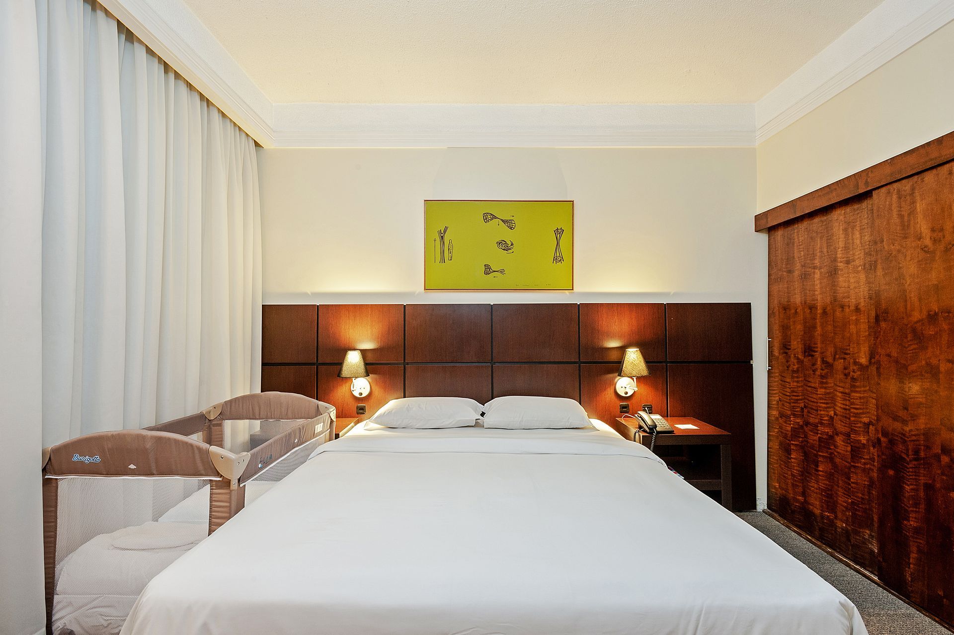 Quarto de hotel com cama grande, cabeceira de madeira, abajures e janela com cortinas.