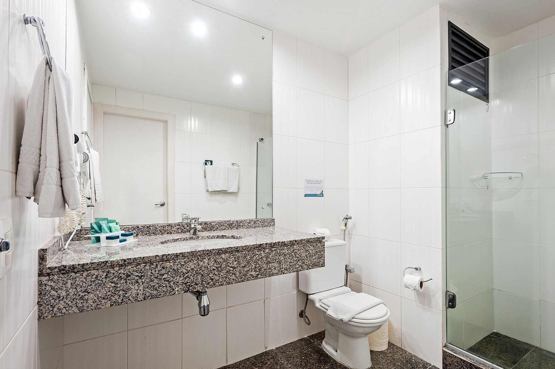 Banheiro moderno e iluminado com espelho de vaidade, bancada de granito, vaso sanitário e box de vidro.