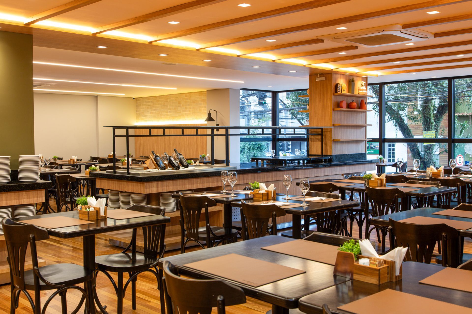 Sala de jantar moderna do restaurante com mesas de madeira, cadeiras pretas e grandes janelas com vista para a vegetação.